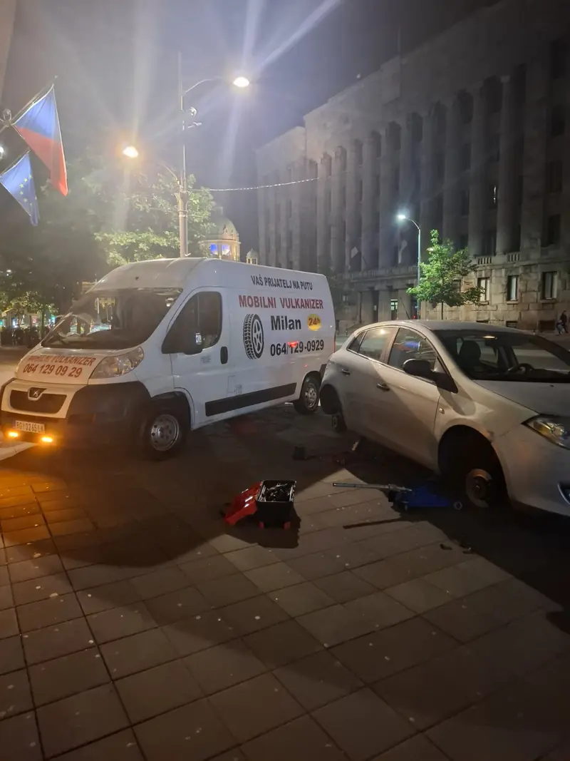 Mobilni vulkanizer Milan noću ispred Skupštine Srbije, servisiranje belog automobila sa hidrauličnom dizalicom