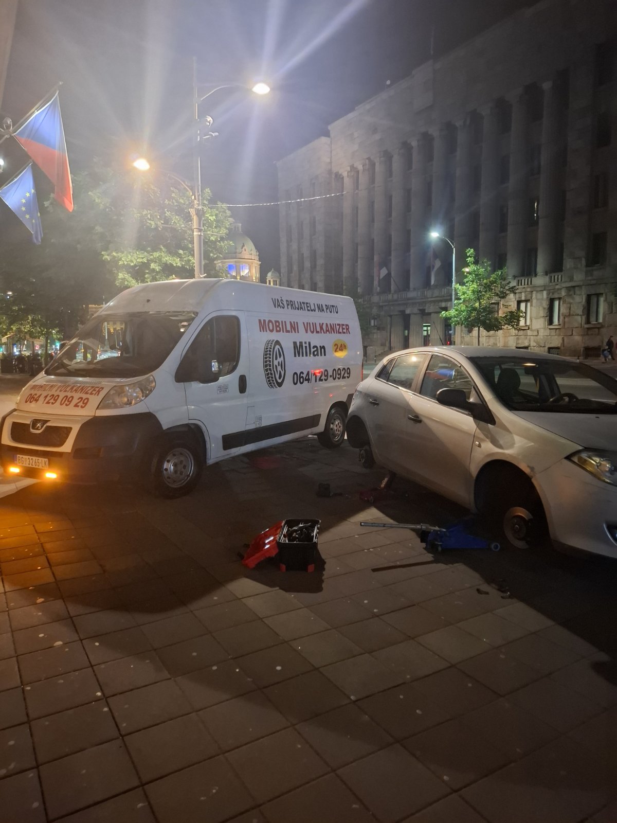 Mobilni vulkanizer Milan noću ispred Skupštine Srbije u centru Beograda — servisiranje belog automobila sa hidrauličnom dizalicom i alatom, 24h non-stop